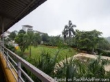 The image shows a view from a balcony of a building. The balcony has a wooden railing and is overlooking a lush green landsca...