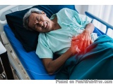 The image shows an elderly man lying in a hospital bed. He is wearing a green hospital gown and has a blue blanket draped ove...