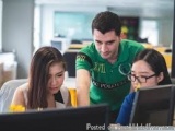 The image shows three young people, two women and a man, sitting in front of a computer monitor in an office setting. The man...