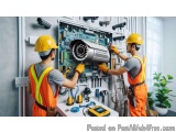 The image shows two men wearing orange safety vests and hard hats, working on a large electrical panel in a room. They are bo...
