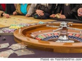 The image shows a group of people sitting around a roulette table in a casino. The table is covered with a purple tablecloth...