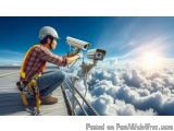 The image shows a construction worker wearing a hard hat and safety vest, standing on a metal platform with a security camera...