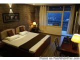 The image shows a bedroom with a large window on the right side. The window has a view of the city and the sky. The room has...