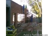 The image shows a construction site where a brick building is being built. There are two workers on a scaffolding platform, w...