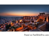 The image is a panoramic view of a city at sunset. The sky is a beautiful orange and pink color, with the sun setting in the...