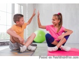 The image shows a boy and a girl sitting on a pink yoga mat in a room with large windows. The boy is wearing an orange t-shir...