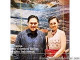 The image shows a man and a woman standing side by side in front of a large mural. The man is wearing a blue traditional Indo...