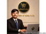 The image shows a man sitting at a desk with a laptop in front of him. He is wearing a black suit and a blue tie. He has a mu...