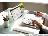 The image shows a person's hands holding a pen and writing on a piece of paper while sitting at a desk with a laptop, a calcu...