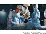 The image shows two surgeons performing an operation in an operating room. They are both wearing surgical gowns, masks, and h...