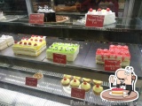 The image shows a display case in a bakery. The display case is filled with different types of cakes and pastries. On the lef...
