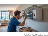 In this image, we can see a man working on an air conditioning unit in a living room. He is wearing a blue uniform and glasse...