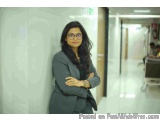 The image shows a young woman standing in a hospital corridor with her arms crossed. She is wearing a grey blazer and glasses...