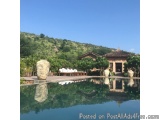 The image shows a beautiful outdoor swimming pool with a thatched roof house in the background. The pool is surrounded by lus...