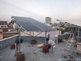 The image shows two men working on a rooftop solar panel installation. The rooftop is located on a flat roof and is surrounde...