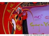 The image shows a person dressed in a traditional Chinese costume, standing on a stage with a red background and gold accents...
