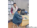 The image shows two men in an office setting. The man on the left is standing behind the desk and is wearing a white vest and...