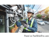 The image shows a young man wearing a hard hat and safety vest, holding a tablet in his hand and smiling at the camera. He is...