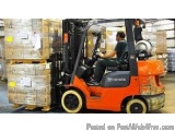 The image shows a man driving a Toyota forklift in a warehouse. The forklift is orange in color and has a black cab with the...