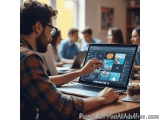The image shows a young man sitting at a wooden table with a laptop in front of him. He is wearing a plaid shirt and glasses...
