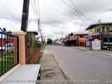 The image shows a street view of a residential area in Trinidad and Tobago. The street is empty and there are several buildin...