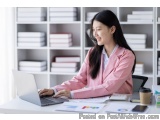 The image shows a young woman sitting at a desk in an office. She is wearing a pink blazer and has long dark hair. She appear...