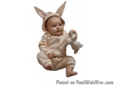 The image is of a baby sitting on the floor and holding a stuffed animal. The baby is wearing a light pink onesie with a hood...