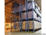 The image shows a large warehouse with a high ceiling and metal beams. The warehouse is filled with rows of blue metal rackin...