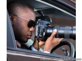 The image shows a man sitting in a car, holding a DSLR camera with a large lens attached to it. He is wearing sunglasses and...