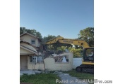 The image shows a yellow Komatsu excavator digging through the rubble of a house. The excavator is digging into the wall of t...