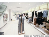 The image shows the interior of a clothing store. The store has a modern and spacious design with white walls and wooden floo...
