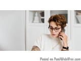 The image shows a young woman with short brown hair and glasses, wearing a white t-shirt. She is sitting in front of a white...