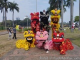 The image shows a group of four Chinese lion dancers performing in a park. The dancers are dressed in traditional red and yel...