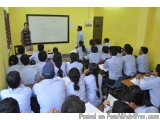 The image shows a classroom with a group of students sitting on benches and facing a whiteboard. The students are wearing blu...