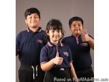 The image shows three young children, two boys and a girl, standing side by side and smiling at the camera. They are all wear...