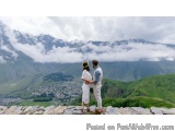 The image shows a couple standing on top of a mountain, looking out at the beautiful landscape below. The couple is standing...