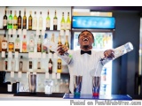 The image shows a bartender at a bar, standing behind the bar counter and pouring a drink from a shaker into a glass. He is w...