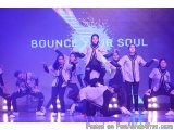 The image shows a group of young dancers performing on a stage. They are wearing matching white and black outfits with the wo...