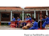 The image shows a group of six men playing musical instruments on a wooden deck. They are all wearing blue traditional Chines...