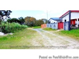 The image shows a dirt road leading to a small white building with a red roof. The building appears to be a single-story stru...