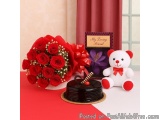 The image shows a table with a bouquet of red roses, a chocolate cake, and a teddy bear. The roses are arranged in a large bo...