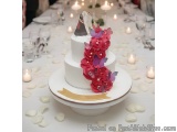 The image is of a three-tiered wedding cake on a white cake stand. The cake is decorated with pink and purple flowers and but...