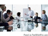 The image shows a group of business people sitting around a conference table in a meeting room. There are six people in total...