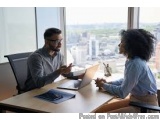 The image shows a man and a woman sitting at a desk in an office, engaged in a conversation. The man is on the left side of t...