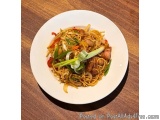 The image shows a plate of noodles with meat and vegetables on a wooden table. The noodles are yellow and appear to be stir-f...