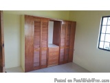 The image shows a wooden wardrobe in a room with a window on the right side. The wardrobe is made of wood and has multiple do...