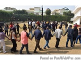 The image shows a group of people holding hands and walking in a circle in a park. The people are of different ages and ethni...