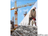 The image shows a construction site with a large yellow tower crane in the background. The crane is lifting a large piece of...