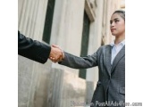 The image shows two people shaking hands in front of a building. The person on the left is a man wearing a black suit and tie...