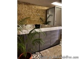 The image shows a modern bathroom with a large bathtub in the center. The bathtub is made of gray tiles and has a white count...